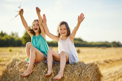 Two charming girls are sitting on a roll of mown rye in a field in summer