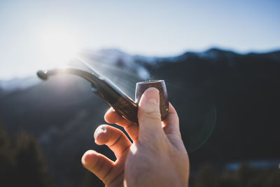 Cropped hand of man holding pipe cigarette against sky