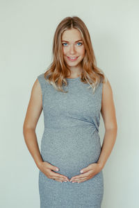 Portrait of pregnant young woman touching abdomen while standing against white background