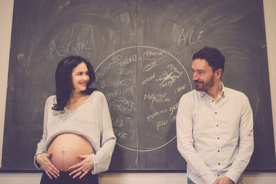 Smiling pregnant woman looking at man against blackboard