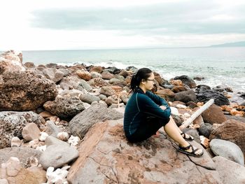 Woman sitting on rock at beach against sky