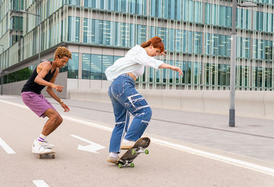 Full body of happy multiracial friends in casual wear riding skateboards on asphalt roadway while having fun together in city