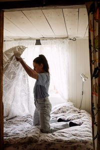 Side view of young woman folding blanket while kneeling on bed in cottage