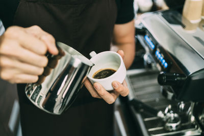 Crop image of a young male barista pouring hot milk into espresso black coffee for making latte art.