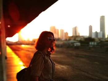 Side view of young woman standing against cityscape at sunset