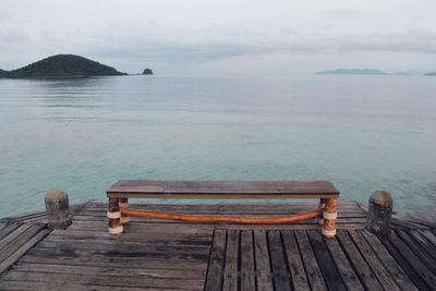 Wooden bench on pier by sea against sky