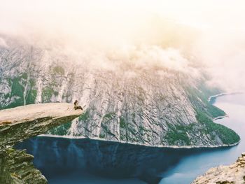 Hiker sitting on cliff enjoying view of mountain and river against cloudy sky during foggy weather