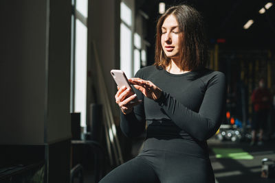 Portrait of resting young brunette woman in sport active wear using mobile in fitness club