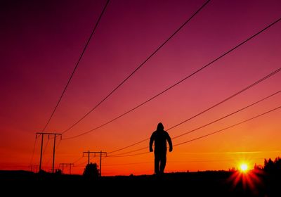 Silhouette man walking against electricity pylon during sunset