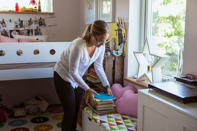 Woman stacking books on table while cleaning bedroom at home