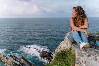 Young woman sitting on rock by sea against sky