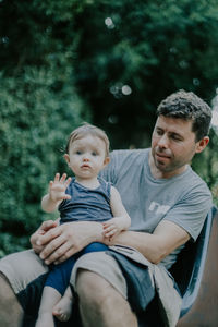 Portrait of caucasian dad with baby girl in his arms showing hello hand gesture sitting on a chair