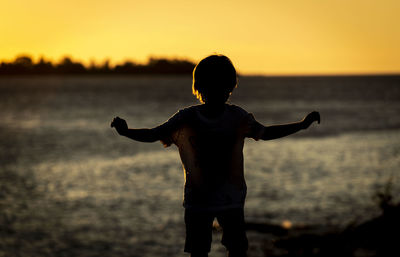 Rear view of silhouette boy standing at beach during sunset