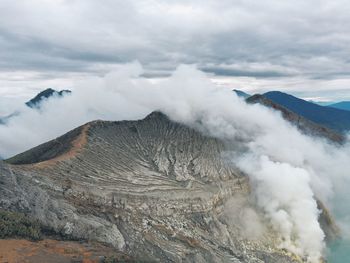 Smoke covering rocky mountains against cloudy sky