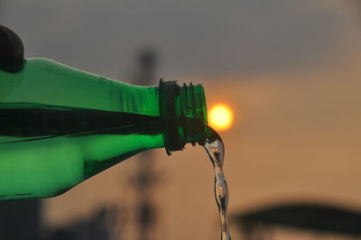 Close-up of person holding bottle against sky during sunset