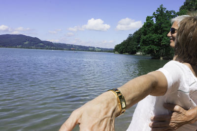 Happy senior couple standing with arms outstretched by lake against sky