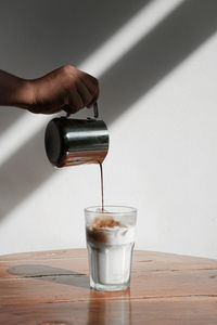 Close-up of hand pouring drink in glass on table against wall