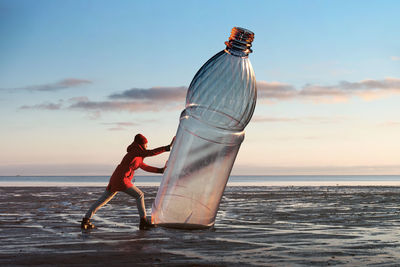 A woman is pushing a huge plastic bottle on the shore.