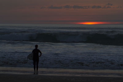 Silhouette man standing with surfboard on shore at beach during sunset