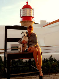 Full length portrait of smiling woman with dalmatian sitting on wooden chair against lighthouse during sunset