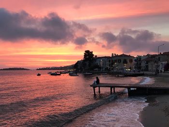 Woman sitting on pier at beach against sky during sunset