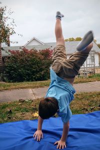 Boy practicing handstand on blue fabric at grassy field against sky