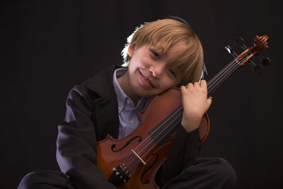 Boy holding violin while sitting against black background