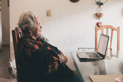 Side view of elderly female with short gray hair sitting on chair making video call via netbook at home