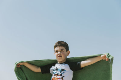 Low angle view of boy holding green towel while standing against clear sky during sunny day