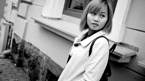 Tilt image of young woman with backpack looking away while standing by building