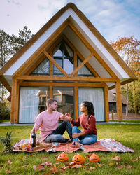 Full length of man and woman sitting on yard with wine and jack o lantern on grass 
