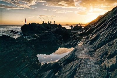 Scenic view of fisherman at sea on rocks against cloudy sky