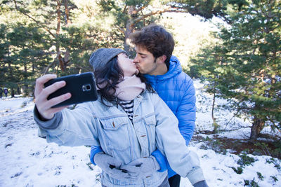 Young couple kissing while taking selfie with mobile phone during winter