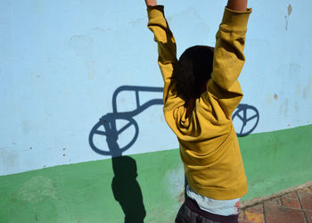 Rear view of girl standing with arms raised by wall