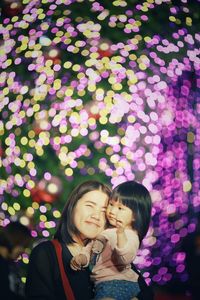 Portrait of smiling woman carrying daughter against illuminated lights at night