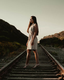 Side view of young woman standing on railroad track against clear sky