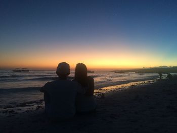 Rear view of couple sitting at beach against sky during sunset