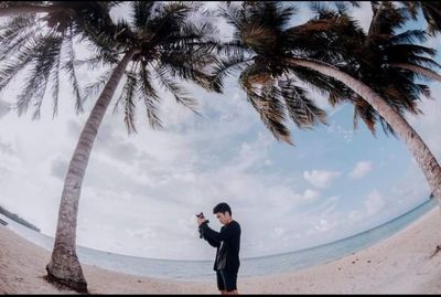 Man photographing palm trees on beach against sky