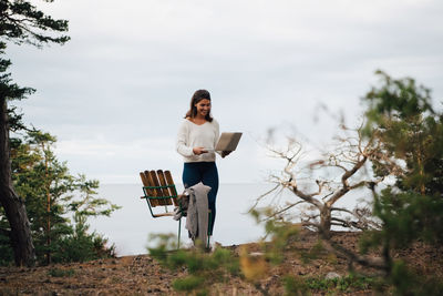 Smiling young woman with laptop standing by chair in forest against sky