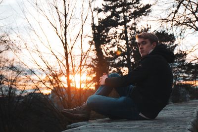 Portrait of man sitting on retaining wall against trees during sunset