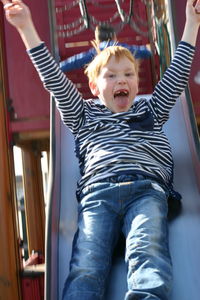 Portrait of cheerful boy sliding with arms raised at playground