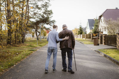 Full length rear view of caretaker supporting retired senior man walking with cane on road