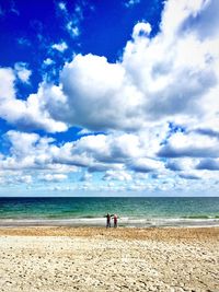 Distant view of friends with arms outstretched standing at beach against cloudy sky