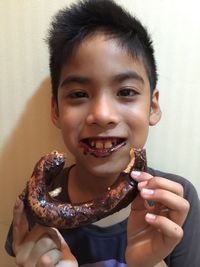 Close-up portrait of happy boy eating chocolate pretzel by wall