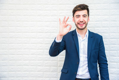 Portrait of smiling businessman showing ok sign while standing by wall