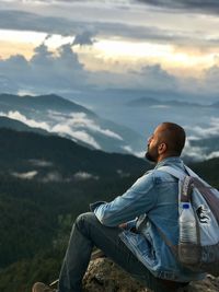 Side view of man with backpack sitting on mountain against sky at sunset