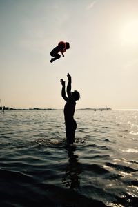 Silhouette shirtless father catching child while standing on sea against sky