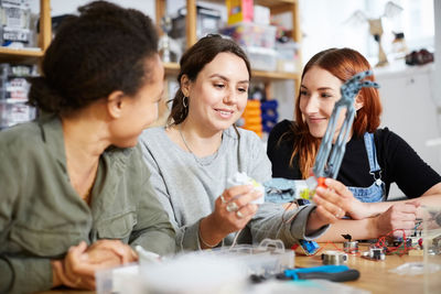 Smiling female inventors sitting with robot model at table in workshop