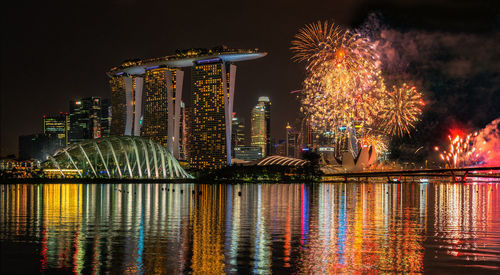 Firework display over river against illuminated buildings in city at night