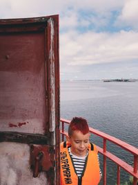 Boy wearing life jacket standing by railing against lake